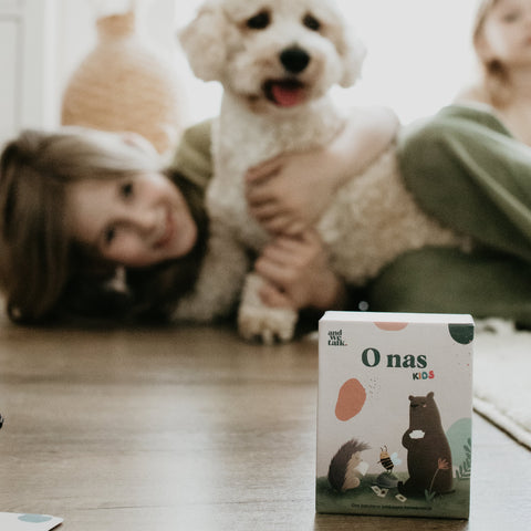 Egy dobozos társasjáték látható előtérben, háttérben egy mosolygó gyerek és egy kutya a padlón